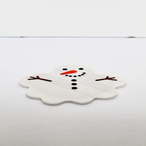 May include: A flat, white, puddle-shaped sticker or decal featuring a melted snowman design. The graphic includes a bright orange carrot nose, black circular eyes, a dotted black mouth, a vertical row of black buttons, and two brown stick arms.