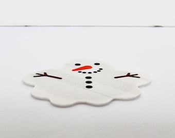 May include: A flat, white, puddle-shaped sticker or decal featuring a melted snowman design. The graphic includes a bright orange carrot nose, black circular eyes, a dotted black mouth, a vertical row of black buttons, and two brown stick arms.