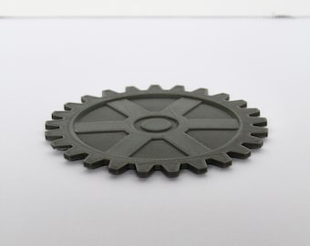 May include: A dark gray, circular gear-shaped object with a central hub and six radiating spokes. The outer edge features evenly spaced teeth, and the surface has a matte finish. The object is positioned flat, showing its top profile clearly.