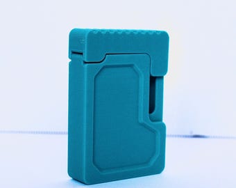 May include: A rectangular teal-colored case with a matte finish and a textured top edge. The front features a recessed panel with beveled edges and a vertical cutout on the right side, suggesting a functional storage container or protective housing.