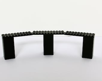 May include: A black construction toy bridge assembly composed of three vertical rectangular support pillars and three horizontal flat plates with stud connectors on top, arranged in a slightly curved, connected linear formation.