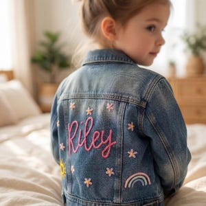 Peut inclure: Une veste en jean bleu avec le prénom Riley brodé en écriture rose au dos. La veste est décorée de petites fleurs roses brodées, d'un soleil jaune et d'un arc-en-ciel pastel. Le modèle présente un col et des manches longues.