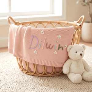 Könnte beinhalten: Eine rosa gestrickte Babydecke mit dem Namen "Dilwin" und Blumenstickereien. Sie liegt in einem Weidenkorb, neben einem weißen Teddybären, was eine gemütliche Szene für das Kinderzimmer schafft.