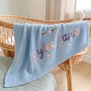 Könnte beinhalten: Eine hellblaue Strickdecke für Babys mit dem eingestickten Namen Rylee Jane in rosa und lila Schreibschrift. Die Decke ist mit kleinen gestickten Bienen und Blumen verziert und liegt über einer hellbraunen Weidenwiege.