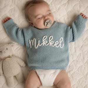 May include: A light blue long-sleeve ribbed knit sweater featuring the name Mikkel embroidered in white cursive script across the chest. The sweater is worn by a sleeping infant with a pacifier, accompanied by a small beige plush lamb toy.