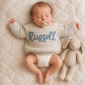 Può includere: Un neonato che dorme indossa un maglione in maglia color crema con macchioline e il nome Russell ricamato in corsivo blu sul petto. Il neonato ha un ciuccio beige in bocca ed è sdraiato accanto a un coniglietto di peluche color beige.