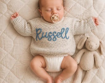 Könnte beinhalten: Ein schlafender Säugling trägt einen cremefarbenen, gesprenkelten Strickpullover mit dem Namen Russell, der in blauer Schreibschrift auf die Brust gestickt ist. Der Säugling hat einen beigen Schnuller im Mund und liegt neben einem beigen Plüschhasen.