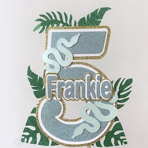 May include: A cake topper featuring the number 5 in glittery blue and gold, with the name "Frankie". It's decorated with light green snakes and tropical leaves. A wooden stick is attached to the bottom.