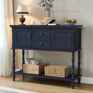 May include: A navy blue wooden sideboard with two doors, two drawers, and a lower shelf. Two woven baskets sit on the shelf. A lamp, vase, and bowl of fruit are on top. The text "HearthWooden" is visible.