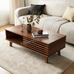 May include: A rectangular, dark brown wooden coffee table with a slatted drawer front and open shelf. The table has four angled legs and is placed on a light-colored rug. Decorative items, including a vase and books, are on the table. The text "HearthWood" is visible.