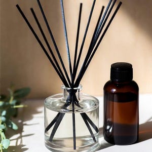May include: A clear glass diffuser bottle with black reeds and a small amber glass bottle with a black cap. The diffuser bottle contains a clear liquid. The background is a neutral beige colour.