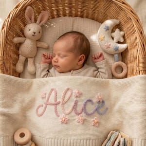 Può includere: Un neonato che dorme in una cesta di vimini, coperto da una coperta in maglia color crema con il nome Alice ricamato in filo rosa e viola con piccoli fiori. Un coniglietto di peluche e un sonaglio a forma di mezzaluna sono vicini.