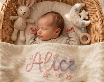 May include: A sleeping infant in a wicker basket, covered by a cream-colored knit blanket featuring the name Alice embroidered in pink and purple yarn with small flower accents. A plush rabbit toy and a crescent moon rattle are placed nearby.