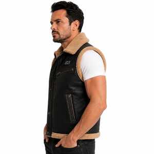 May include: A black sleeveless vest with a tan faux shearling collar and trim. The vest features a front zipper closure, two zippered side pockets, and a small rectangular patch on the chest with the text B3 printed in white lettering.