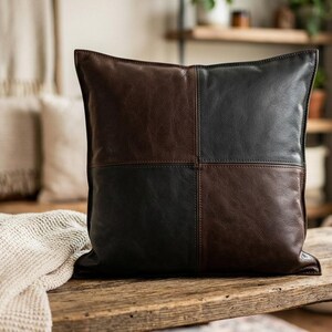 May include: A square throw pillow featuring a color-blocked design with four alternating panels of dark brown and black leather. The pillow has visible stitching along the central vertical and horizontal seams, creating a distinct grid pattern.