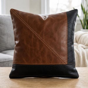 May include: A square throw pillow featuring a color-blocked design with a large brown section and a black vertical panel on the right side, separated by a diagonal seam. The pillow has a black horizontal band along the bottom edge with stitching.