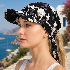 May include: A black sun hat featuring a white floral pattern with light gray leaves. The hat has a wide, structured brim at the front and a gathered crown that tapers toward the back, secured with two long fabric ties hanging down the side.