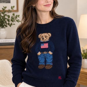 May include: A navy blue long-sleeve knit sweater featuring a central graphic of a teddy bear wearing a sweater with an American flag design, blue jeans, and brown shoes. The letters RL are embroidered in red on the lower left side of the sweater.
