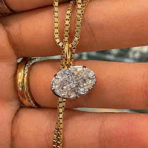May include: A gold box chain necklace featuring a single oval-shaped pendant set with multiple small, clear, faceted stones. The pendant is held by a small gold bail attached to the chain, which is draped vertically across the fingers.