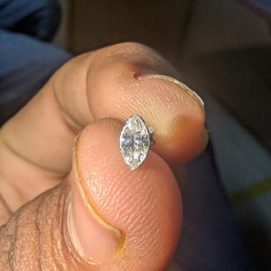 Puede incluir: Un pendiente de botón con un diamante de talla marquesa, que presenta una piedra transparente facetada montada en un engaste de metal plateado pulido. La gema tiene una forma ovalada puntiaguda con múltiples facetas reflectantes.