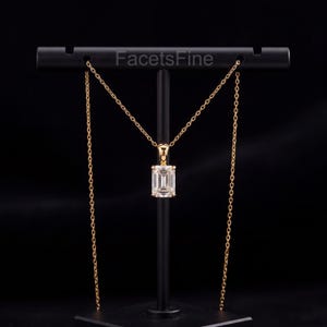Könnte beinhalten: Eine goldfarbene Halskette mit einem klaren, smaragdgeschliffenen Edelsteinanhänger an einer feinen Kette. Der Schmuck wird auf einem schwarzen T-förmigen Ständer präsentiert, auf dessen Querstange der Text FacetsFine weiß aufgedruckt ist.