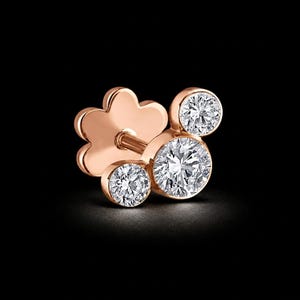 May include: A rose gold stud earring featuring three round, brilliant-cut diamonds. The earring has a flower-shaped backing and a post for insertion. The diamonds sparkle against the dark background, showcasing the jewelry's elegance.