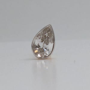 Pode incluir: Uma pedra preciosa transparente e facetada em forma de pera, engastada numa moldura de metal prateado lisa e polida. A pedra apresenta um topo pontiagudo e uma base arredondada, refletindo a luz para um aspeto brilhante e cintilante.