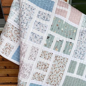 May include: A patchwork quilt with a variety of floral and patterned fabrics in soft pastel colors. The quilt features squares of blue, pink, and green, separated by white borders. The quilt is made of soft, quilted fabric.