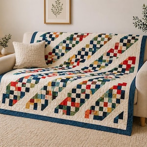 May include: A colorful quilt draped over a cream-colored sofa. The quilt features diagonal stripes of checkered squares in red, blue, green, and yellow against a cream background. A blue border frames the quilt. A beige pillow rests on the sofa.