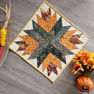 May include: A quilted, star-shaped table topper with a geometric design. The quilt features shades of orange, brown, green, and white. The edges are finished with a yellow border. The quilt is approximately 18 inches square.