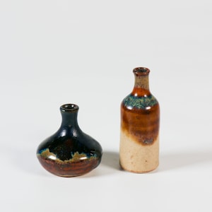 Pode incluir: Dois pequenos vasos de cerâmica com esmalte marrom, azul e branco. O vaso à esquerda é redondo e o vaso à direita é alto e cilíndrico.