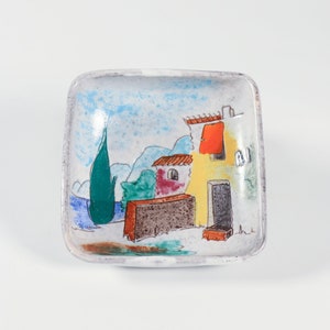 Puede incluir: Un plato cuadrado de cerámica blanca con una escena pintada a mano de una casa amarilla con un techo rojo y un árbol verde en frente. La casa tiene una puerta roja y una ventana. El fondo es un cielo azul con nubes blancas.