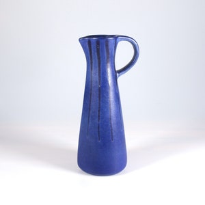 May include: A cobalt blue ceramic jug with a handle. Vertical dark blue lines run down the body of the jug. The jug has a narrow neck and a wide base. The background is plain white.