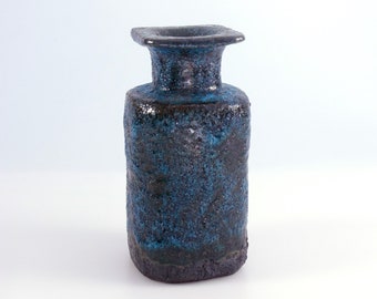 Puede incluir: Un jarrón de cerámica azul oscuro con una abertura cuadrada. El jarrón tiene un cuerpo cilíndrico y un cuello acampanado. El color es una mezcla de azul oscuro y negro, con una superficie rugosa. El jarrón mide unos 20 cm de alto.