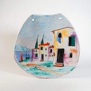 Puede incluir: Colgante de pared de cerámica pintada a mano con una escena de pueblo costero. La obra de arte presenta edificios con detalles en rojo, naranja y morado, veleros y un cielo azul. Dos agujeros en la parte superior para colgar.