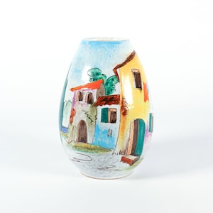 Puede incluir: Un jarrón de cerámica pintado a mano con una escena de pueblo caprichosa. El jarrón presenta un cielo azul, edificios coloridos con tejados rojos y árboles verdes. El jarrón tiene forma redondeada y mide unos 20 cm de alto.