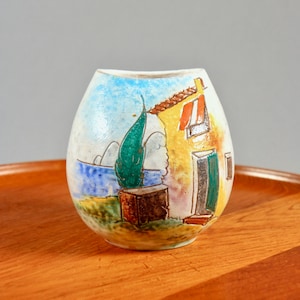 Puede incluir: Un jarrón de cerámica con una escena costera pintada a mano. El jarrón presenta un edificio amarillo con un tejado rojo, un árbol verde y un mar y cielo azules. El jarrón está sobre una bandeja de madera.