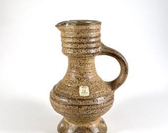 May include: A speckled, brown ceramic pitcher with a handle and spout. The pitcher has a bulbous base and a cylindrical neck with horizontal ridges. A small label is affixed to the front.