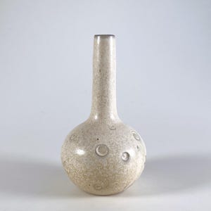 Puede incluir: Un jarrón de cerámica beige con base redonda y cuello largo y delgado. El jarrón tiene una textura moteada y varias hendiduras circulares en la base. La parte superior del cuello es de un tono gris más oscuro.