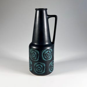 May include: A tall, matte black ceramic pitcher with a single handle and a tapered neck. The lower body features a repeating pattern of teal-coloured, square-shaped motifs with rounded corners, each containing a dark, concentric circular design.