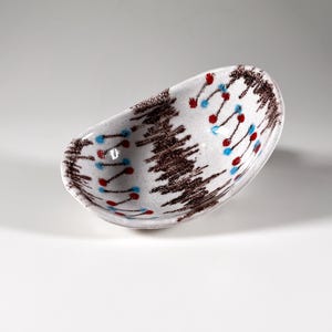 Peut inclure: Bol en céramique de forme ovale, à base blanche. Il est décoré de motifs abstraits bruns, rouges et bleus. Le motif brun ressemble à un arbre, et les points rouges et bleus sont disposés en motif. Le bol est probablement utilisé pour servir des aliments ou comme pièce décorative.