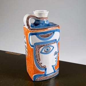 May include: A rectangular ceramic jug featuring a white base with hand-painted abstract designs in orange, blue, and dark brown. The vessel has a single handle at the top, a narrow neck, and a stylized profile of a face depicted on the front surface.