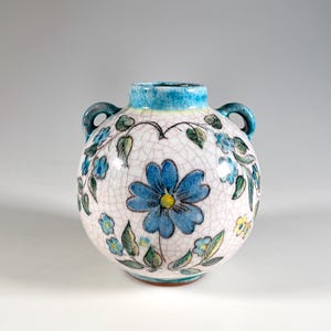 Puede incluir: Un jarrón de cerámica redondo con dos asas, decorado con flores azules y hojas verdes sobre un fondo blanco. El borde y las asas son de color azul turquesa. El jarrón tiene un esmalte craquelado.