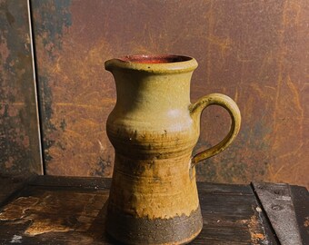 May include: A ceramic pitcher with a brown and yellow glaze. The pitcher has a rounded body and a handle. It is sitting on a wooden surface.