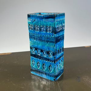 May include: A rectangular ceramic vase featuring a vibrant blue and turquoise glaze. The surface is decorated with horizontal bands of embossed geometric patterns, including small squares, repeating X shapes, and diamond motifs in relief.