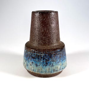 Könnte beinhalten: Eine Keramikvase mit einem strukturierten, gesprenkelten braunen oberen Teil und einem breiteren Sockel mit einem Farbverlauf aus Blau- und Türkistönen. Die Vase hat eine zylindrische Form mit ausgestelltem Boden.
