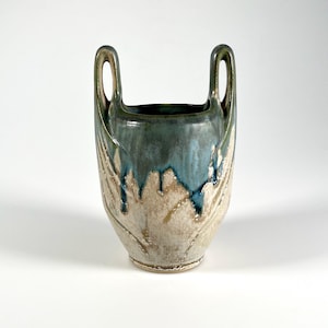 May include: A ceramic vase featuring two tall, integrated handles. The upper portion is glazed in a deep blue-green, which drips down over a textured, light beige base. The vessel has a tapered body and a wide, open circular rim at the top.