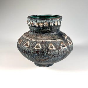 May include: A ceramic vase with a bulbous body and a wide neck, featuring a dark, mottled glaze in shades of blue, black, and brown. The surface has a textured, rugged finish with a row of raised, off-white triangular patterns around the center.
