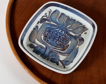 May include: Square ceramic dish with a blue and white floral design. The dish has a white background with a blue and brown floral pattern in the center. The dish is on a wooden surface.