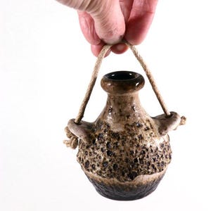 May include: A small, brown and white ceramic vase with a rope handle. The vase has a textured surface and a dark brown glaze.
